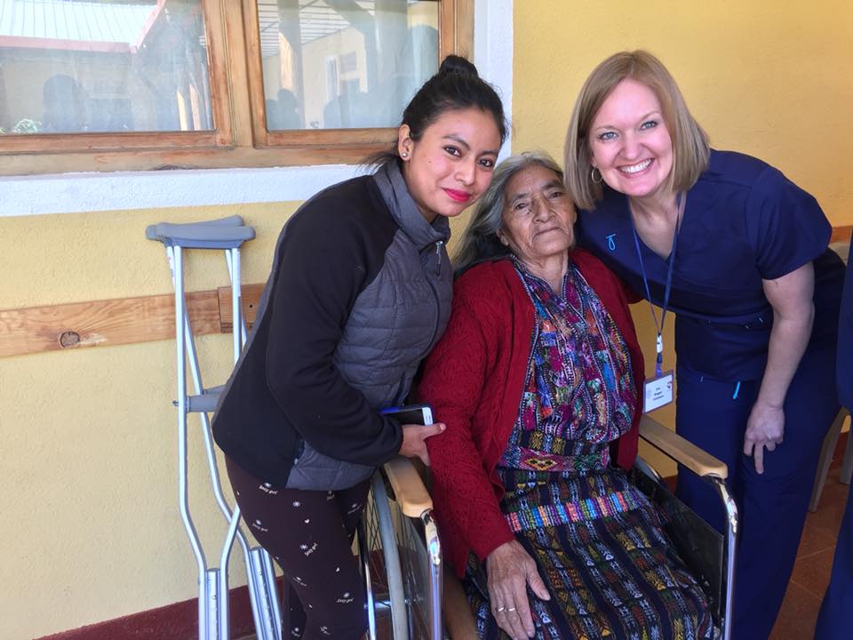 Zwei Frauen und eine ältere Frau im Rollstuhl posieren für ein Foto. Draußen, vor einem Gebäude. Die ältere Frau lächelt.