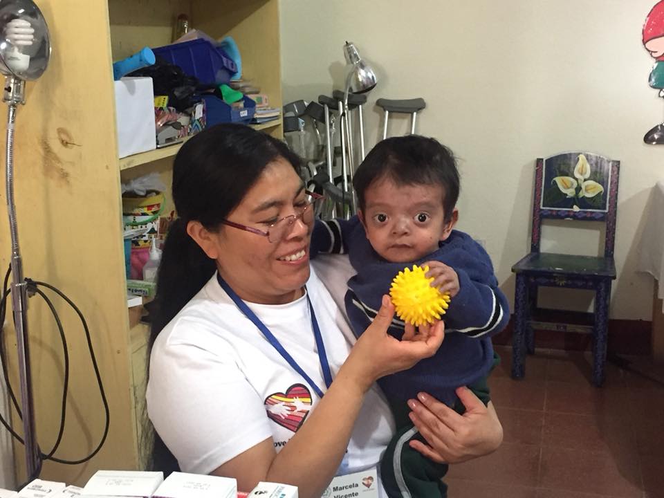 Eine Frau hält ein Kind im Arm, beide lächeln. Das Kind hält einen gelben Ball in der Hand, 
