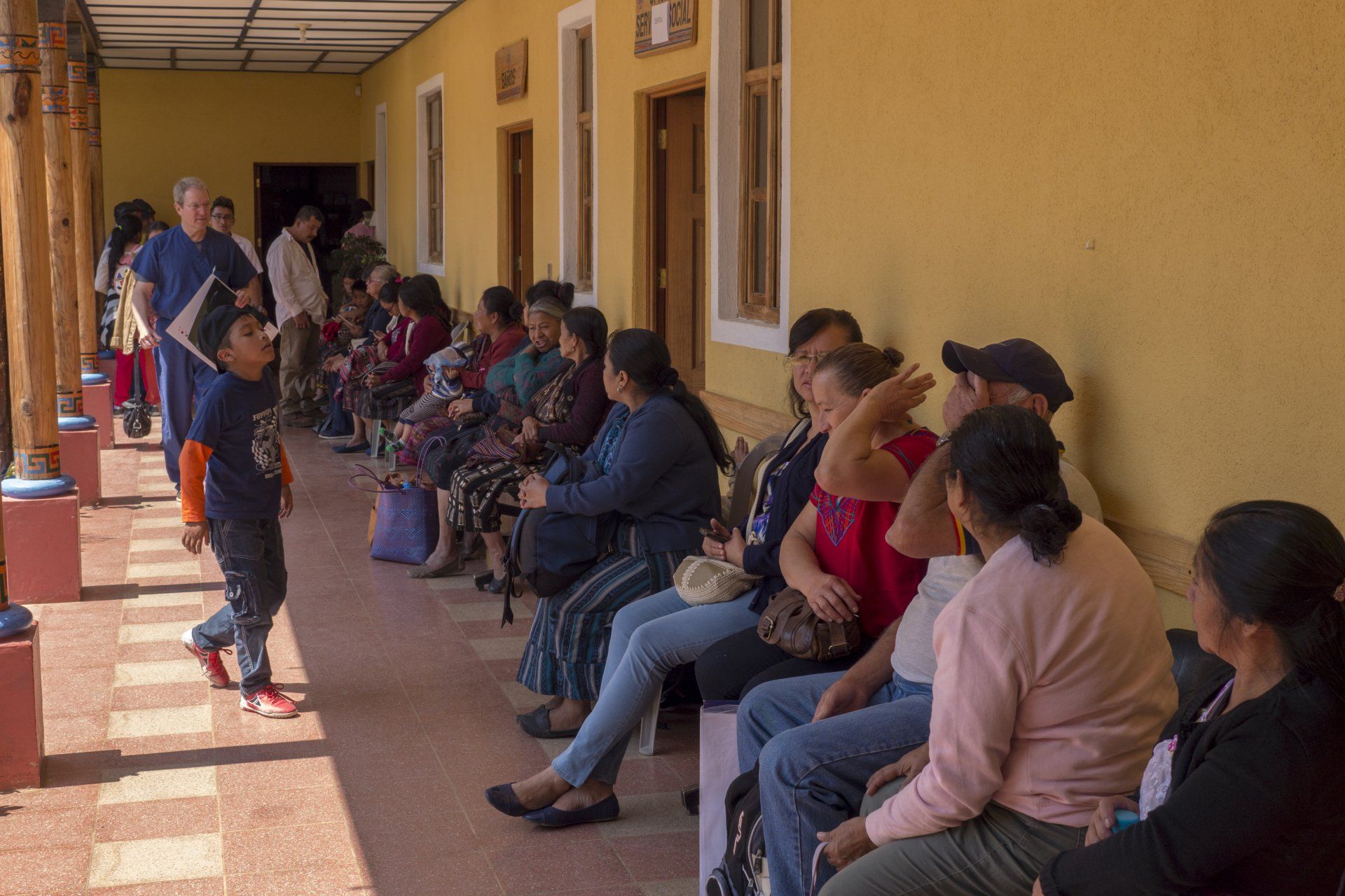 Menschen sitzen auf einer Bank in einem Wartebereich im Freien, einige schauen einem vorbeigehenden Jungen nach.