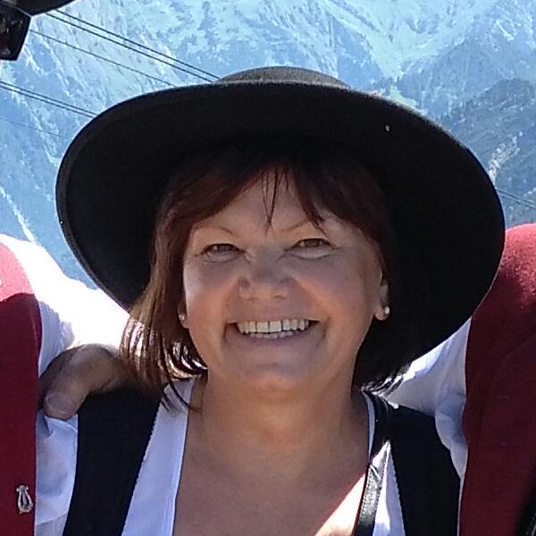 Eine Frau mit schwarzem Hut lächelt vor einer Bergkulisse; im Bild sind auch die Arme einer Person zu sehen.