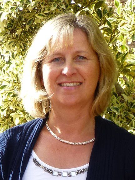 Frau mit blonden Haaren, lächelnd, mit Halskette und dunkelblauem Pullover, vor einem Blätterhintergrund.