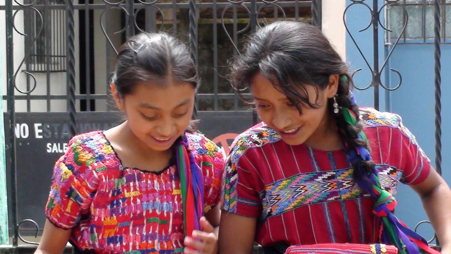 Zwei Mädchen in farbenfrohen, traditionell gewebten Blusen lächeln vor einem Gebäude.