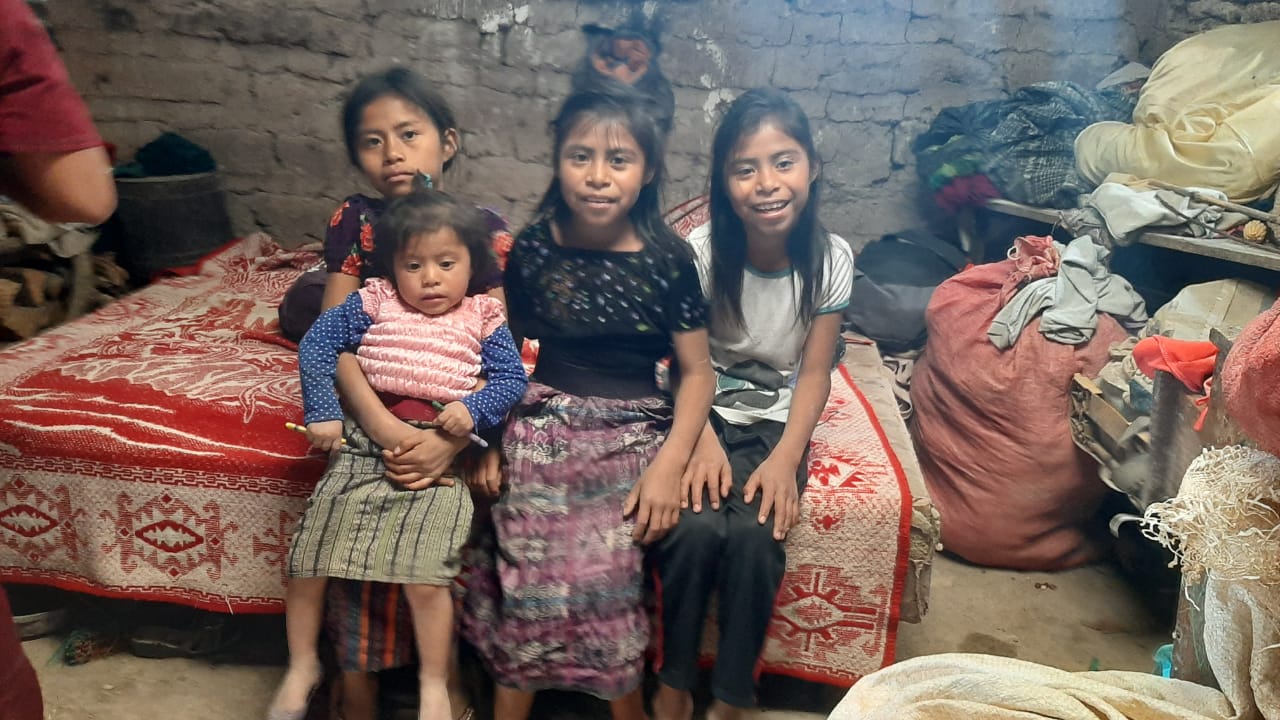 Vier Mädchen und ein Baby sitzen zusammen in einem Haus, in der Nähe eines Bettes und herumliegendem Gerümpel.