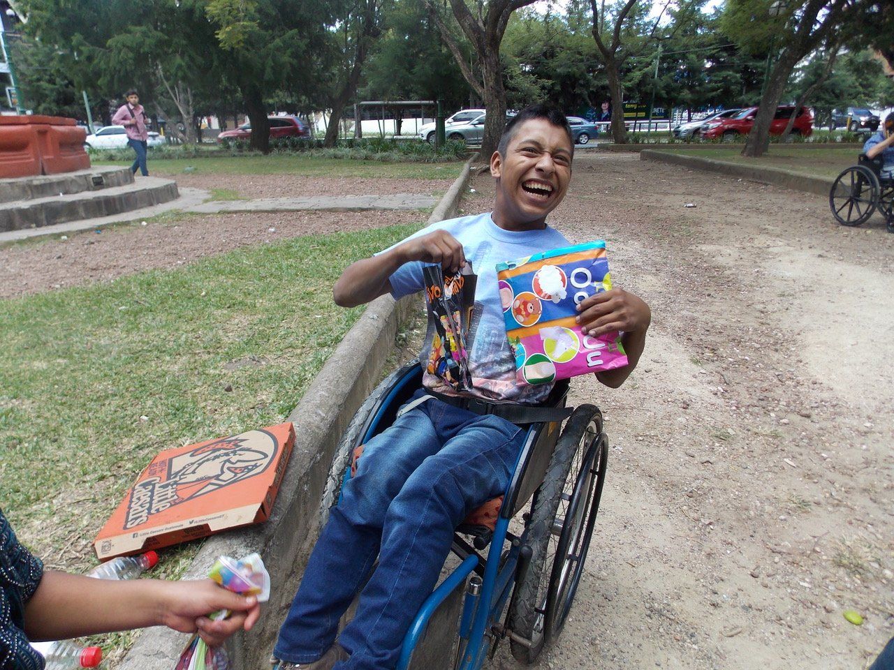 Ein lächelnder Mann im Rollstuhl hält draußen in der Nähe eines Parks Snacks in der Hand.