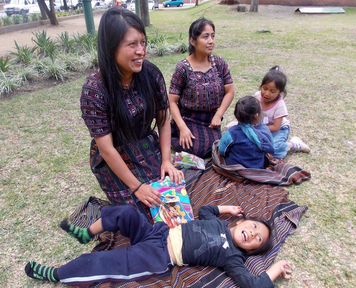 Zwei Frauen und zwei Kinder sitzen lächelnd auf einer Decke in einem Park auf einer Wiese. Ein Kind liegt auf der Decke.
