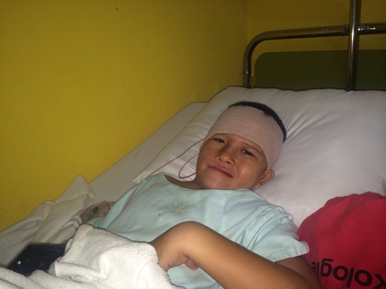 Eine Person mit bandagiertem Kopf liegt im Krankenhausbett, wirkt unbehaglich; neben ihr liegt ein rotes Kissen.