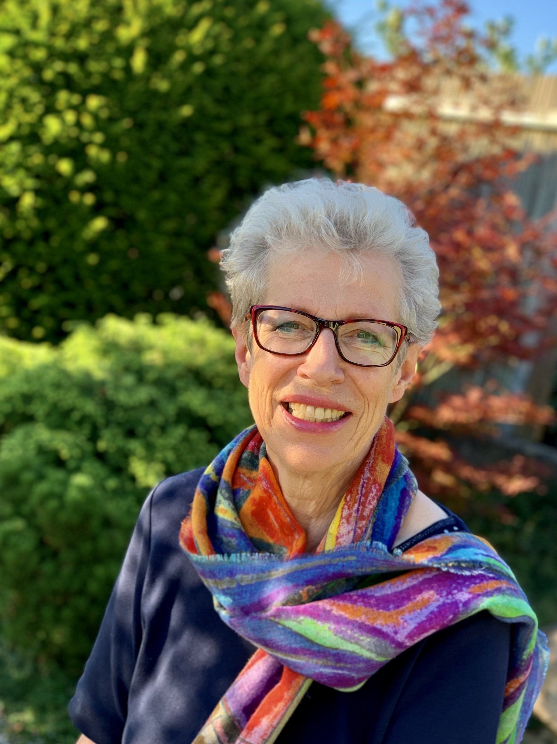 Eine Frau mit kurzem grauem Haar und Brille lächelt im Freien; sie trägt einen bunten Schal.