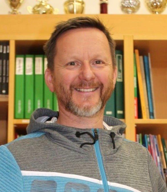 Ein Mann mit Bart lächelt in einem grauen Kapuzenpullover vor Bücherregalen mit Ordnern und Trophäen.