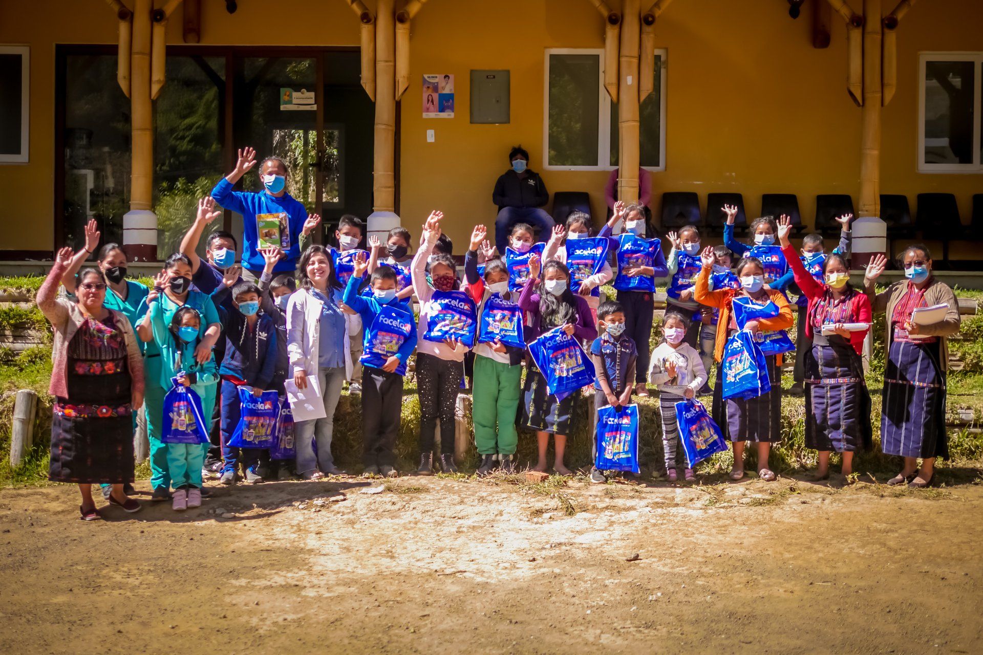 Eine Gruppe von Menschen, viele davon mit Masken, winkt und hält blaue Taschen vor einem Gebäude.