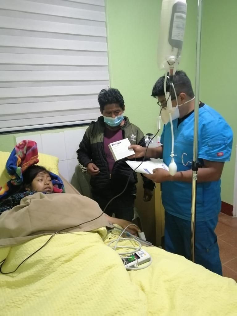 Frau im Bett mit Infusion, Arzt und Mann neben ihr in einem Zimmer, medizinische Geräte sichtbar.