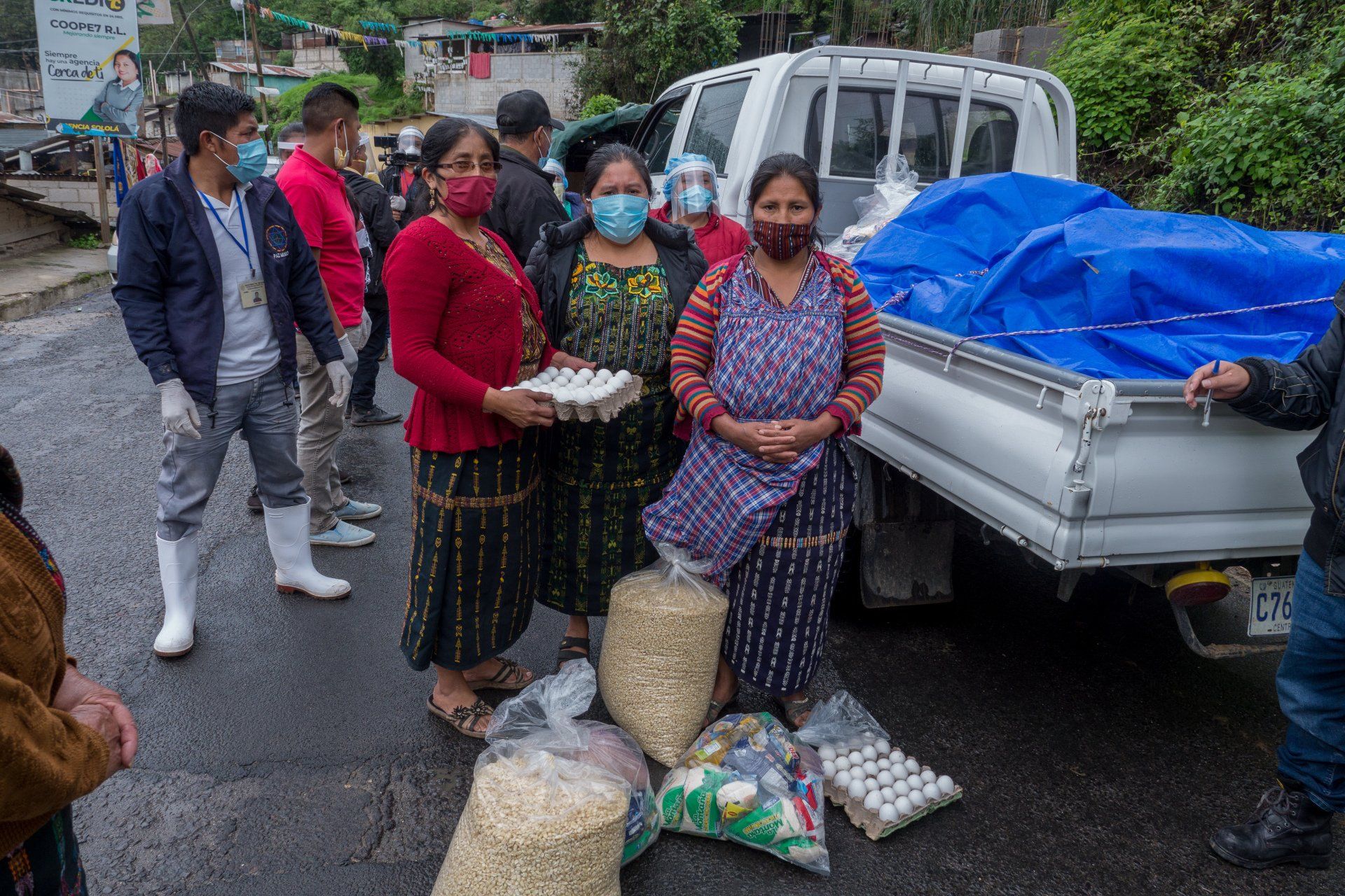Menschen verteilen Lebensmittel, darunter Eier und Säcke mit Getreide, von einem Lastwagen aus. Alle tragen im Freien Masken.