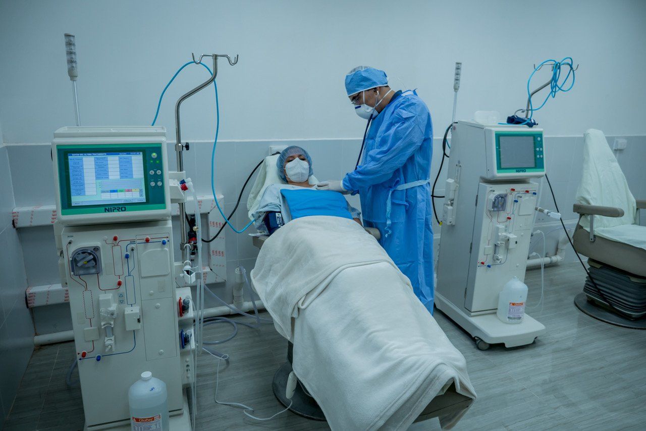 Eine medizinische Fachkraft betreut einen Dialysepatienten; blaue medizinische Geräte, weißer Raum.