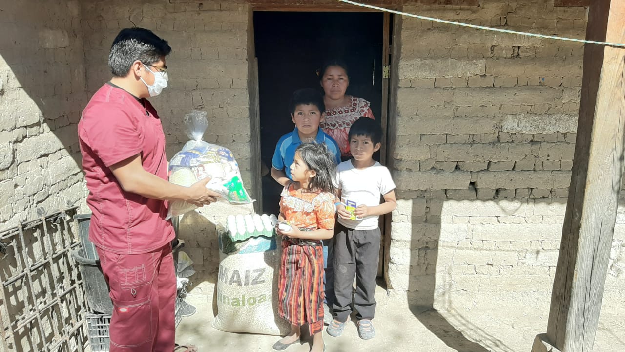 Eine maskierte Person übergibt einer im Türrahmen stehenden Familie Tüten mit Lebensmitteln.
