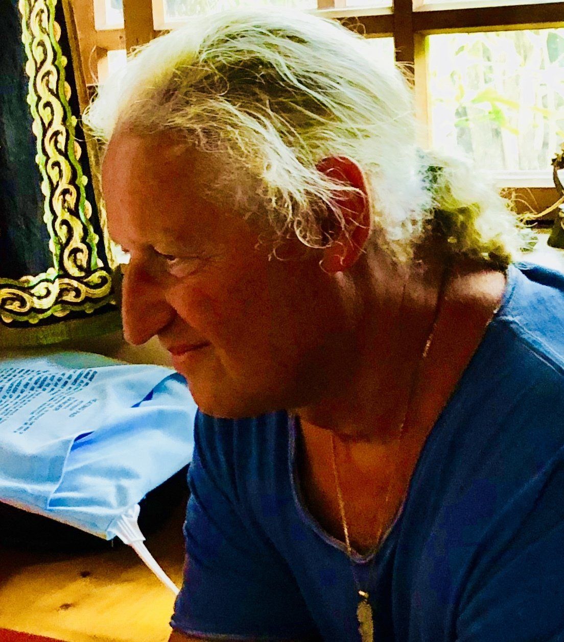 Mann mit langen weißen Haaren in einem blauen Hemd, der nach unten schaut, drinnen in der Nähe eines Fensters.