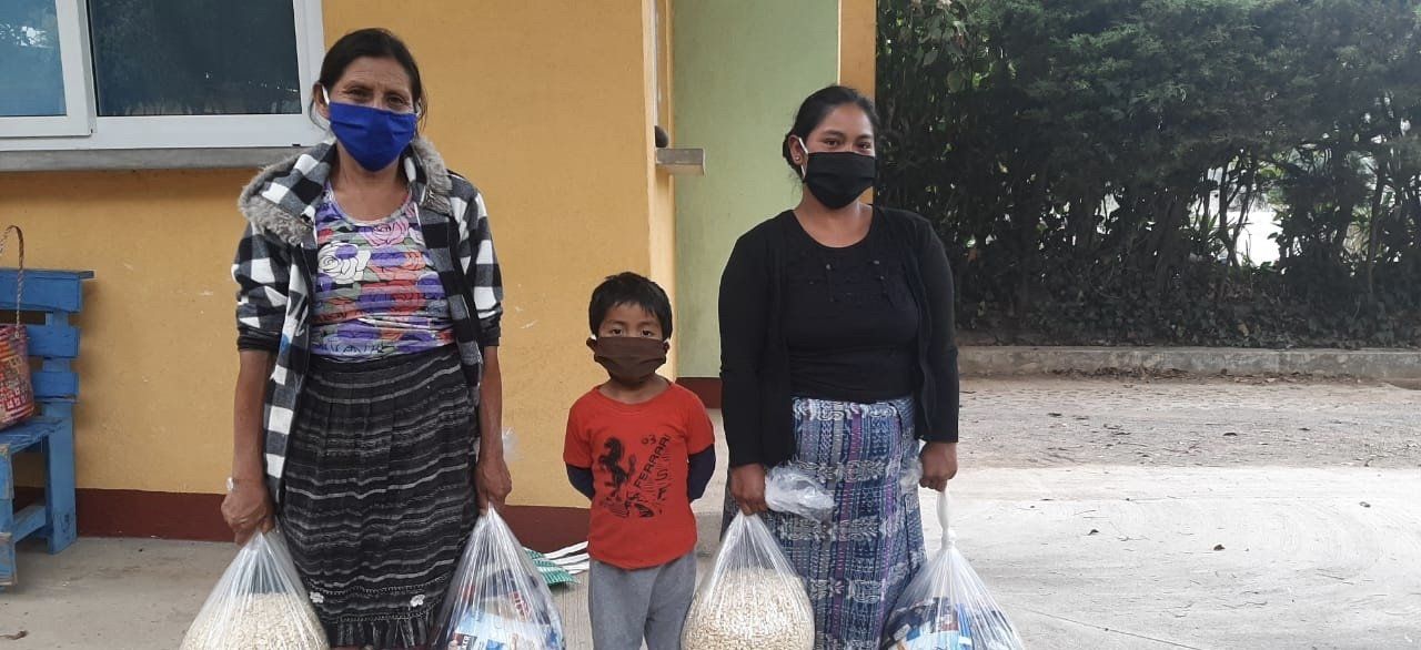 Drei maskierte Personen halten vor einem Gebäude Taschen mit Vorräten.