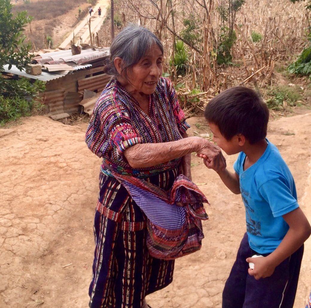 Eine ältere Person in farbenfroher, gewebter Kleidung schüttelt in ländlicher Umgebung einem Kind die Hand.