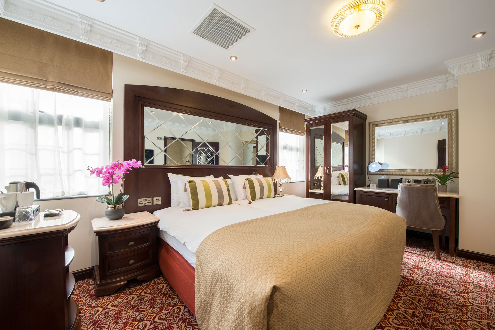 Hotel bedroom with a bed, desk, and wardrobe. Windows on the left, ornate ceiling molding.