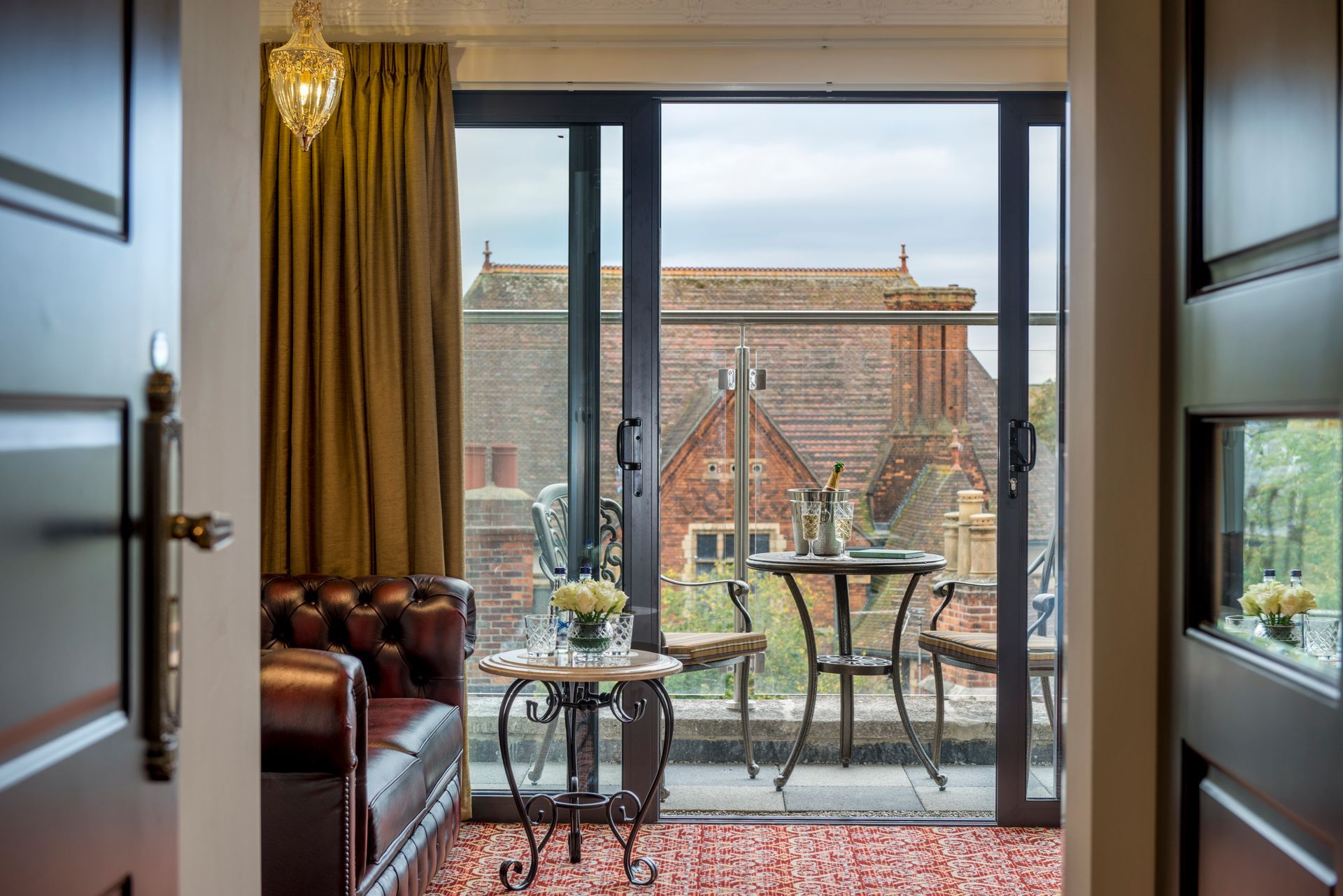 Hotel room with balcony overlooking a building with red tile roof. Brown leather sofa, small table with drinks.