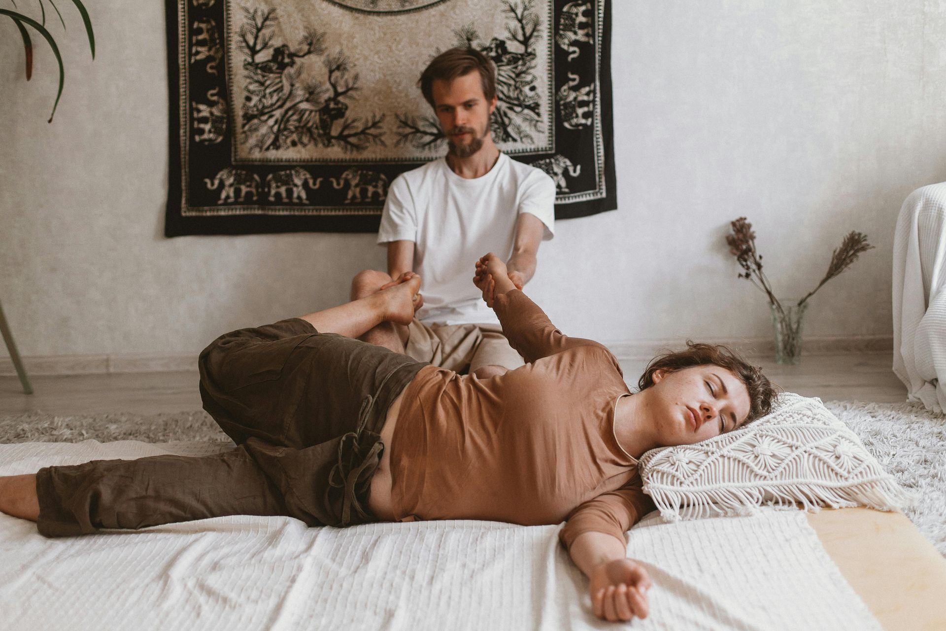 Man giving a woman a Thai massage, working on her leg. Beige room, tapestry, white and brown clothing.