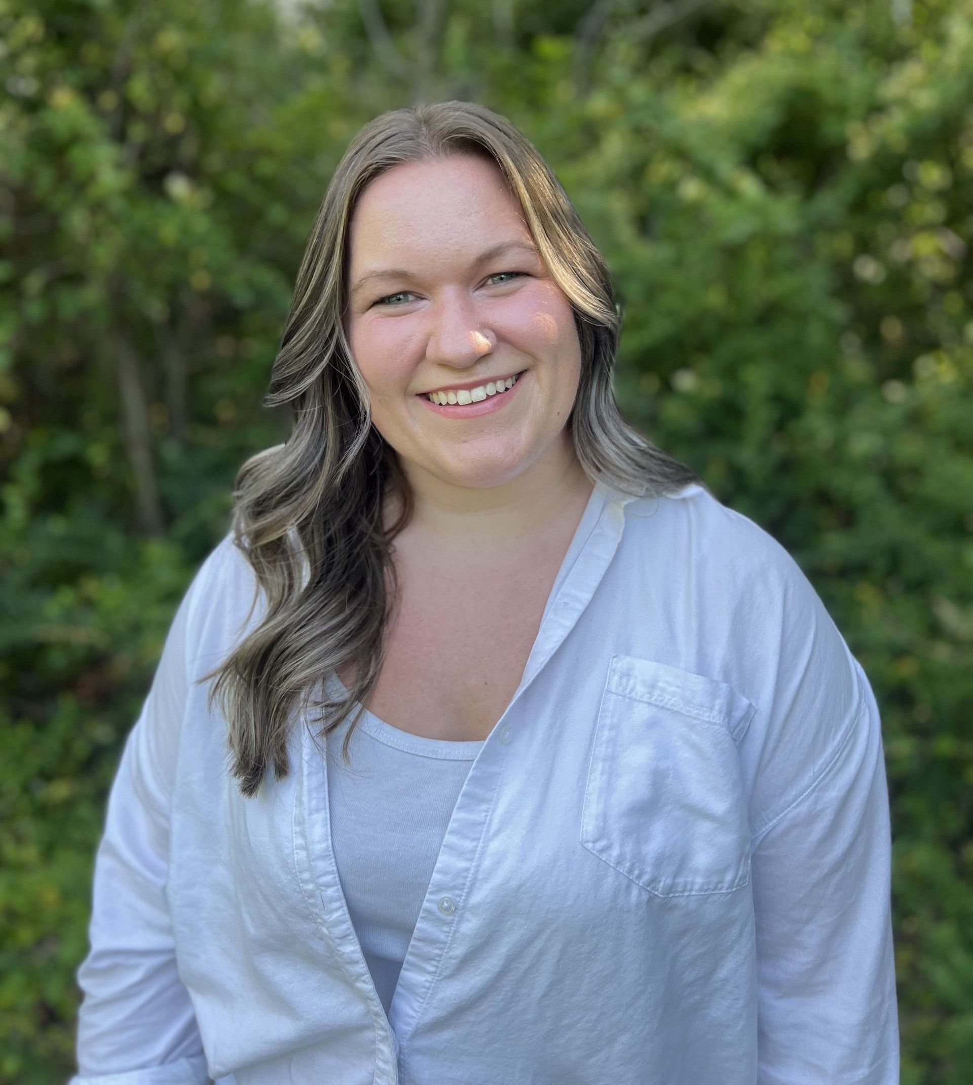 A woman in a white shirt is smiling in front of a forest.