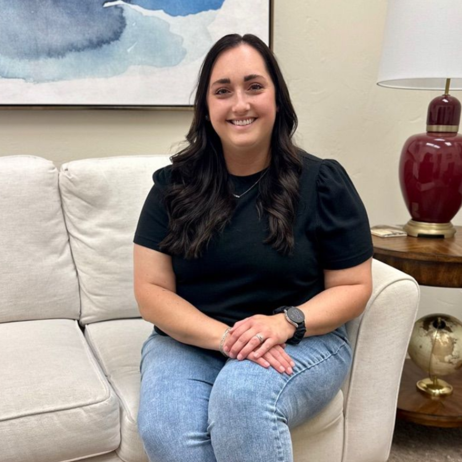 A smiling person with long dark hair, wearing a black shirt and blue jeans, sits on a couch in an office setting.