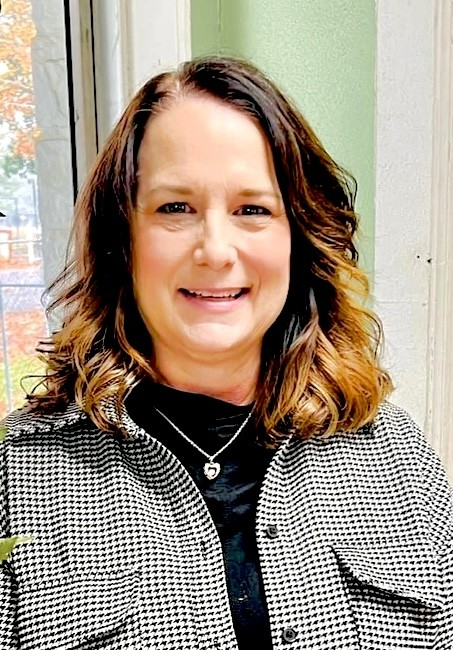 Smiling woman in a black top and patterned jacket standing indoors by a window