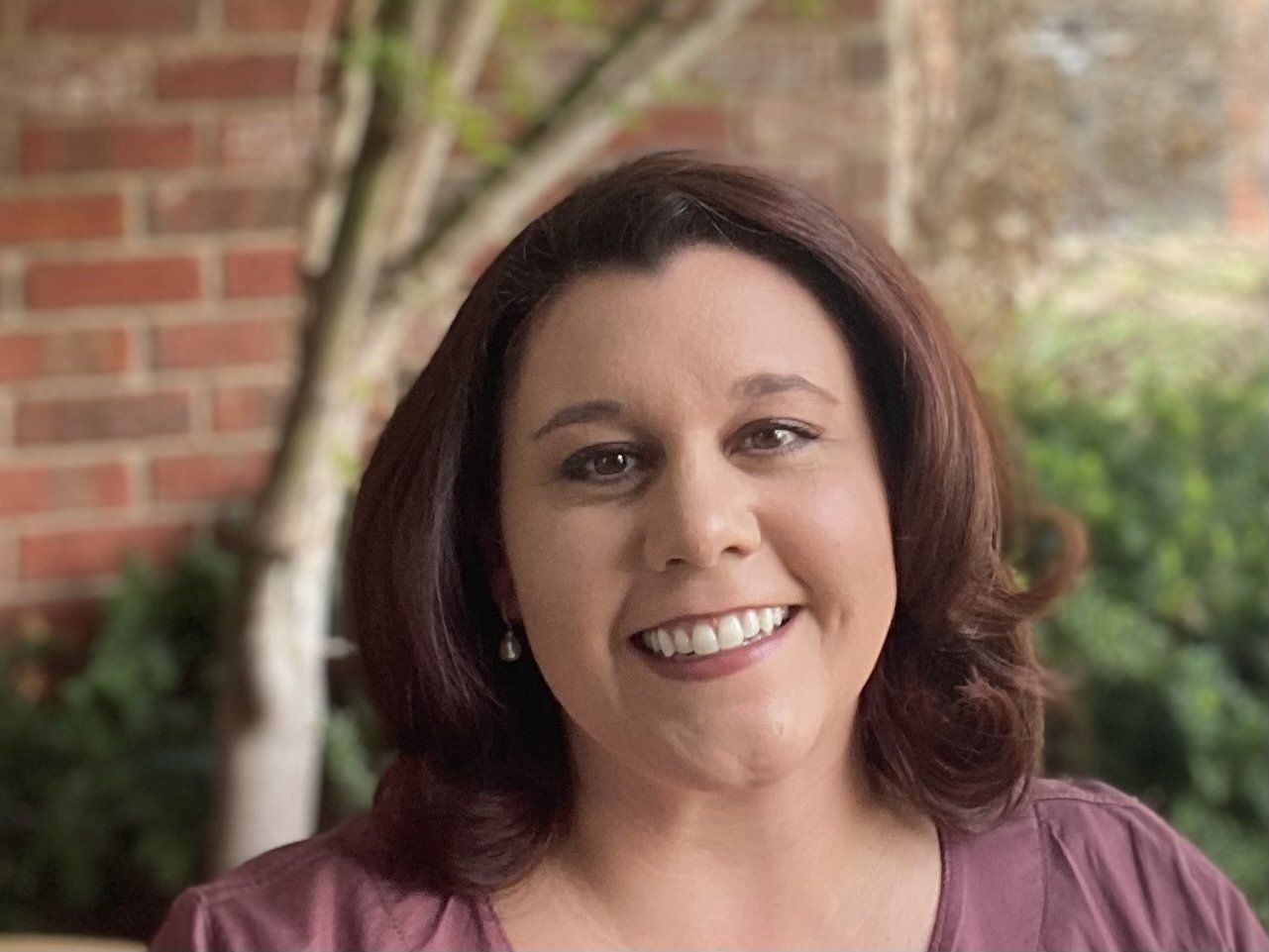 A woman in a purple shirt is smiling in front of a brick wall.