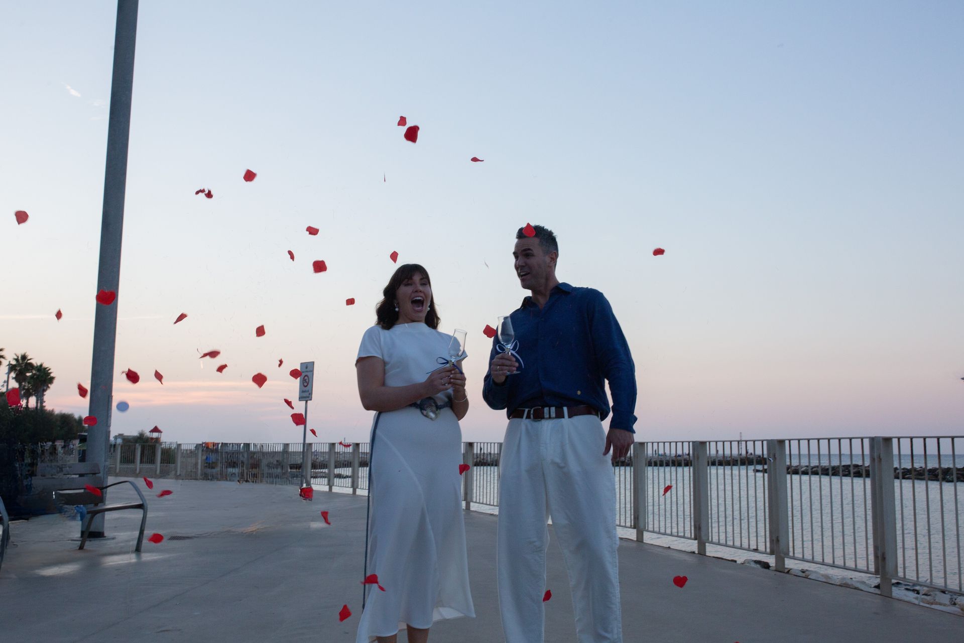 Coppia che festeggia, petali di rosa che cadono. Donna in abito bianco, uomo in camicia blu, champagne, lungomare.