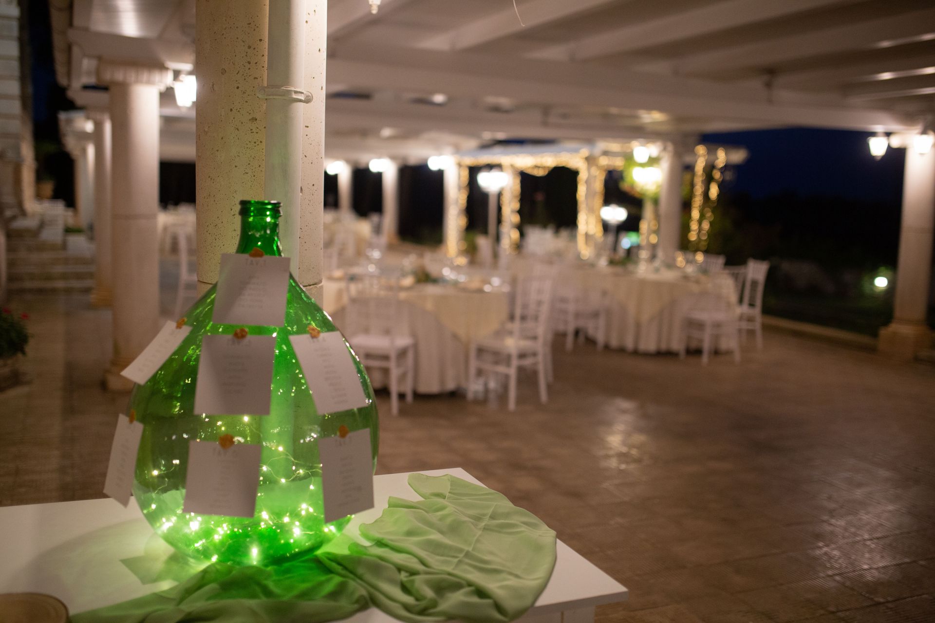Disposizione dei posti a sedere con bottiglie verdi e luci, ricevimento di nozze con tavoli e sedie bianchi al tramonto.