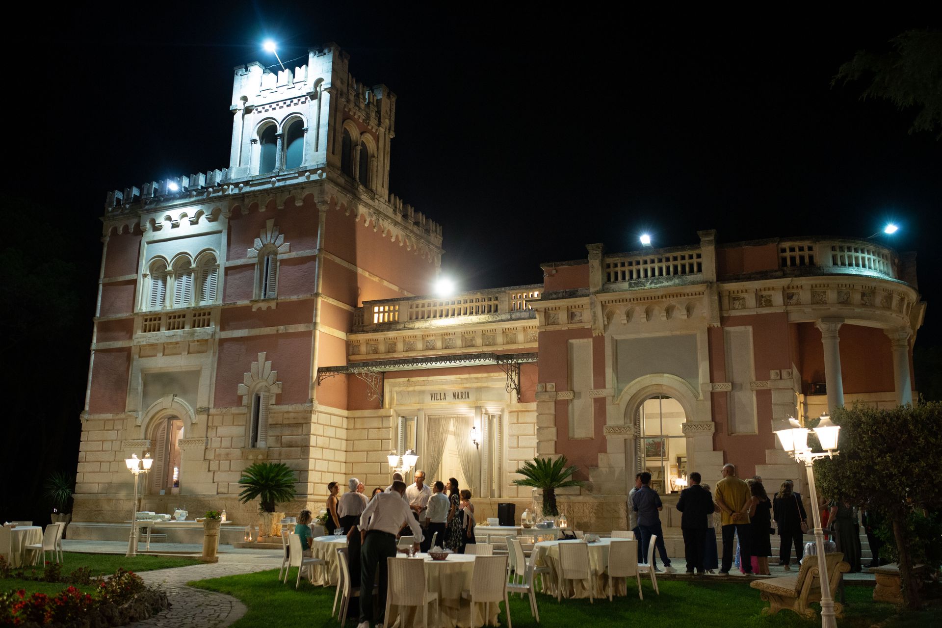 Esterno notturno di un castello rosa con persone sedute ai tavoli sul prato, illuminato da luci.