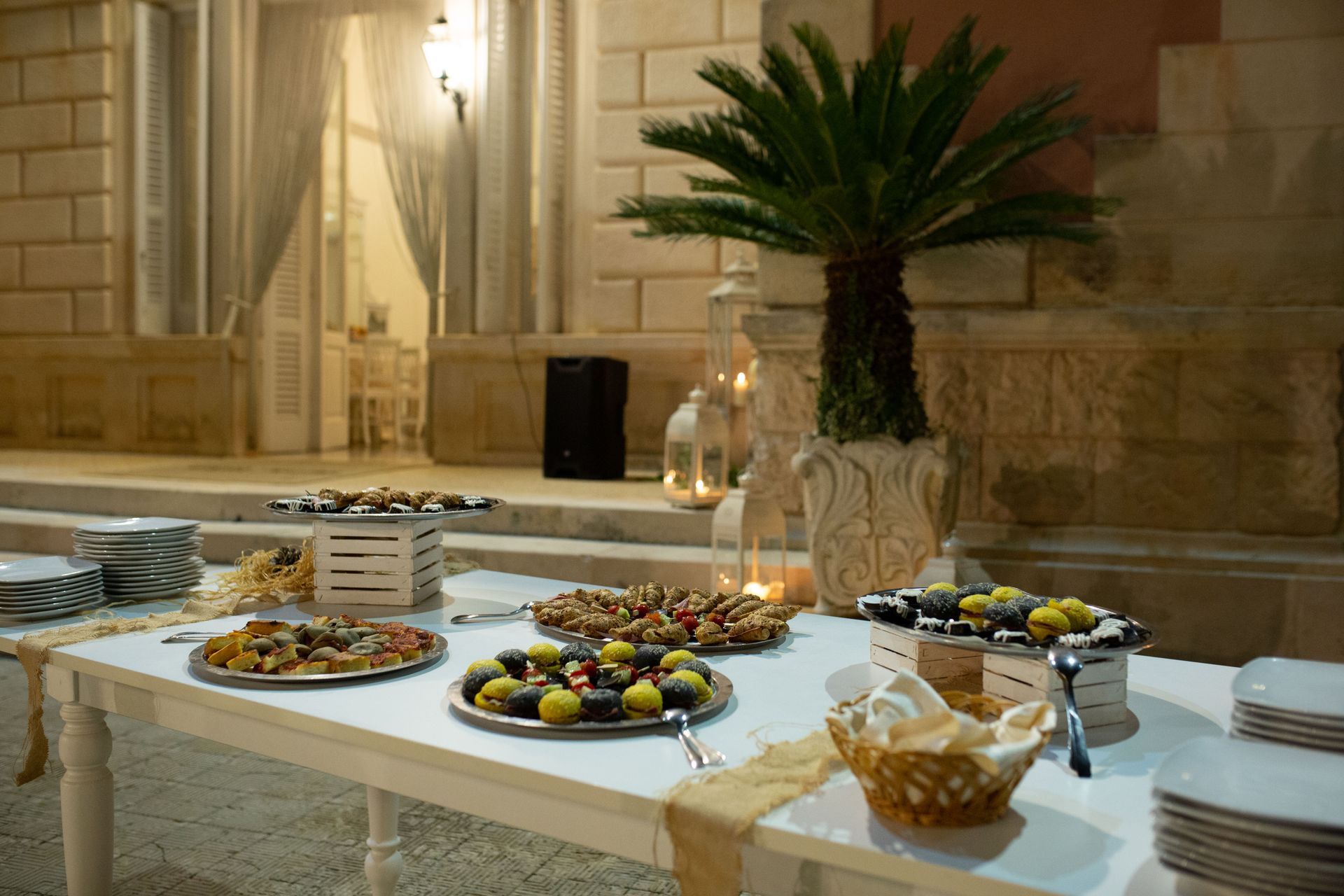 Cibo disposto su un tavolo bianco all'aperto di notte, con una palma, candele e un edificio in pietra sullo sfondo.