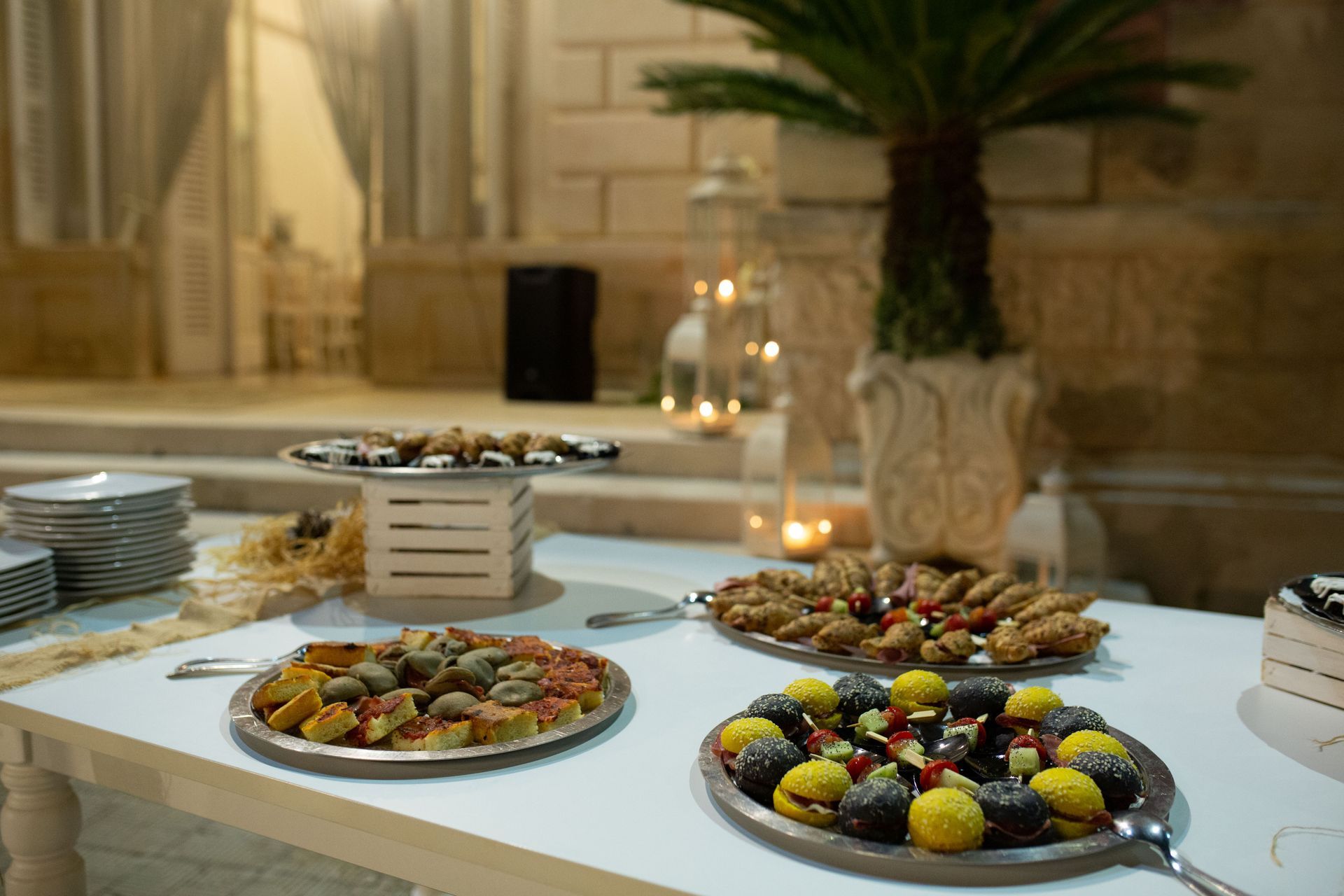 Un tavolo da dessert con pasticcini e dolcetti assortiti, all'aperto durante un evento, con candele e una palma.