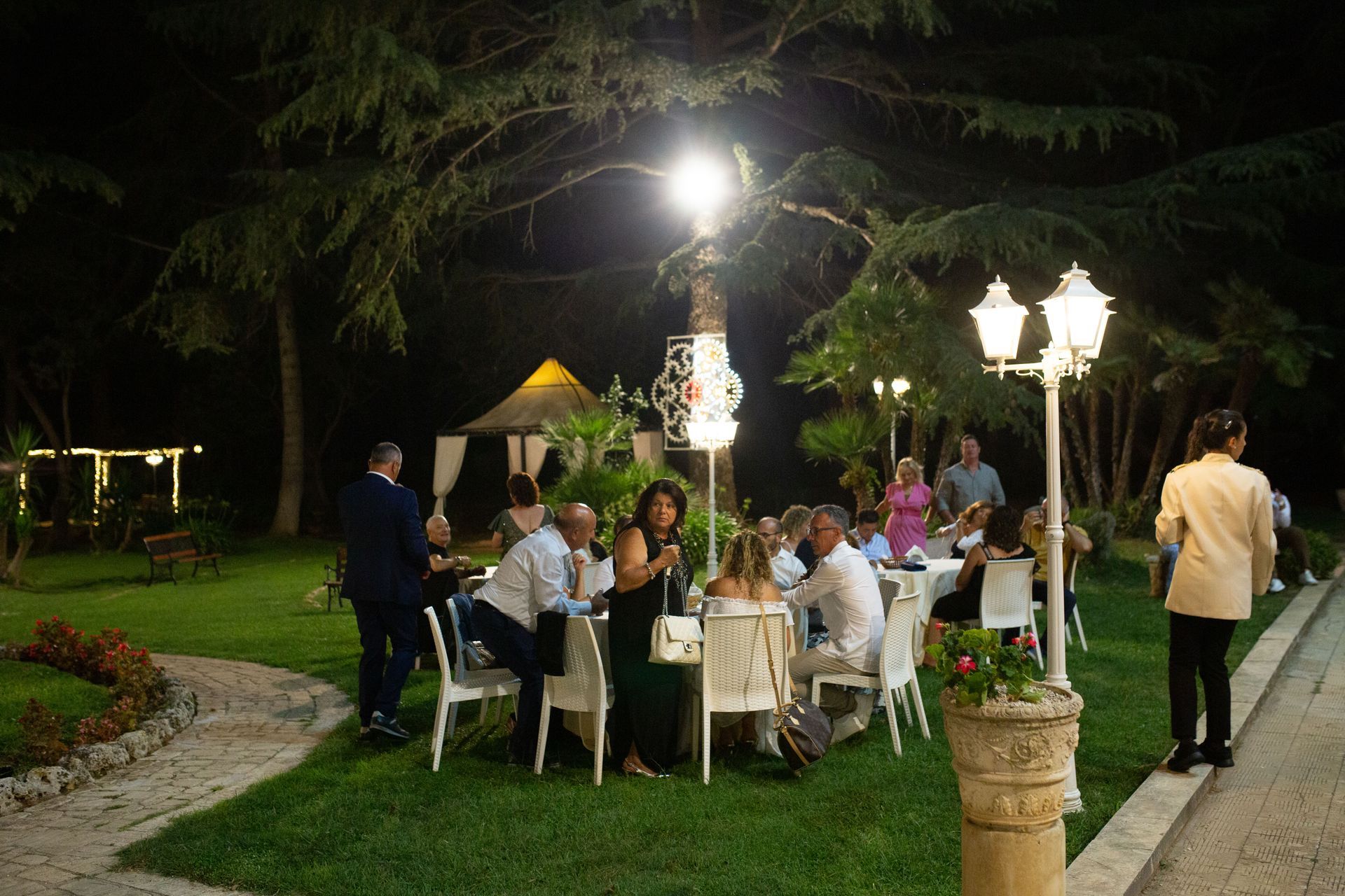 Cena serale all'aperto con un gruppo di persone attorno a un tavolo. Prato verde, sedie bianche e lucine.