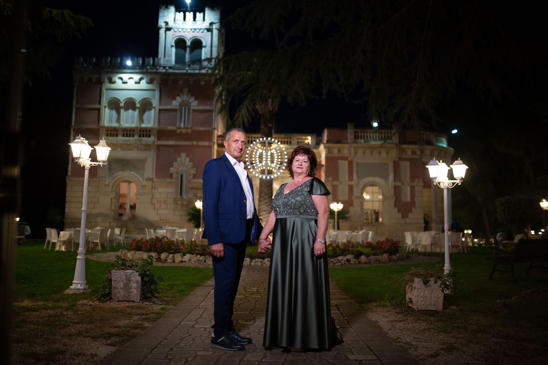 Una coppia si tiene per mano, in posa davanti a un edificio decorato illuminato di notte. Sono in piedi su un sentiero di mattoni, alberi e lampioni incorniciano l'inquadratura.