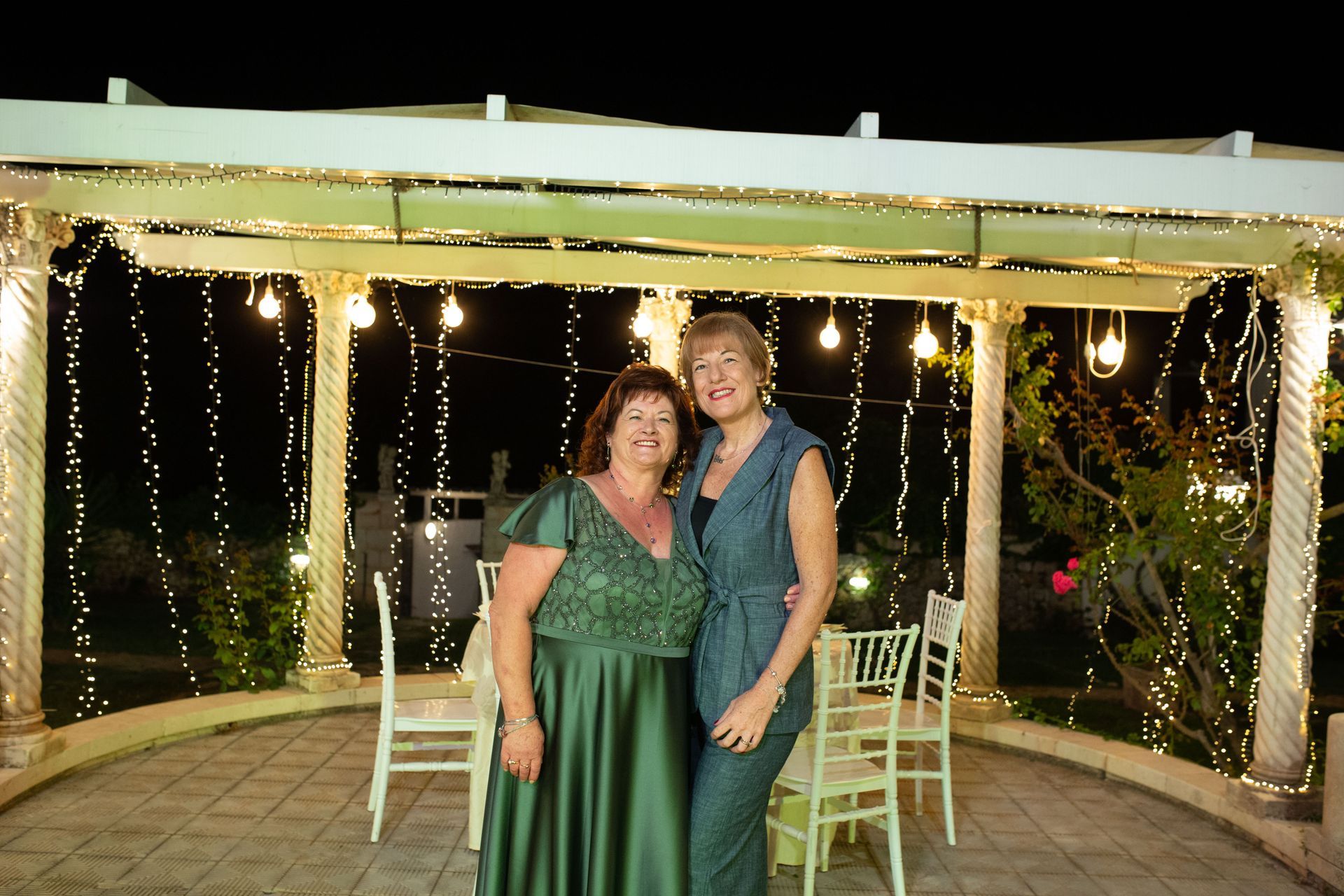 Due donne sono in piedi sotto un pergolato con luci a filo, una con un vestito verde e l'altra con un tailleur pantalone blu.