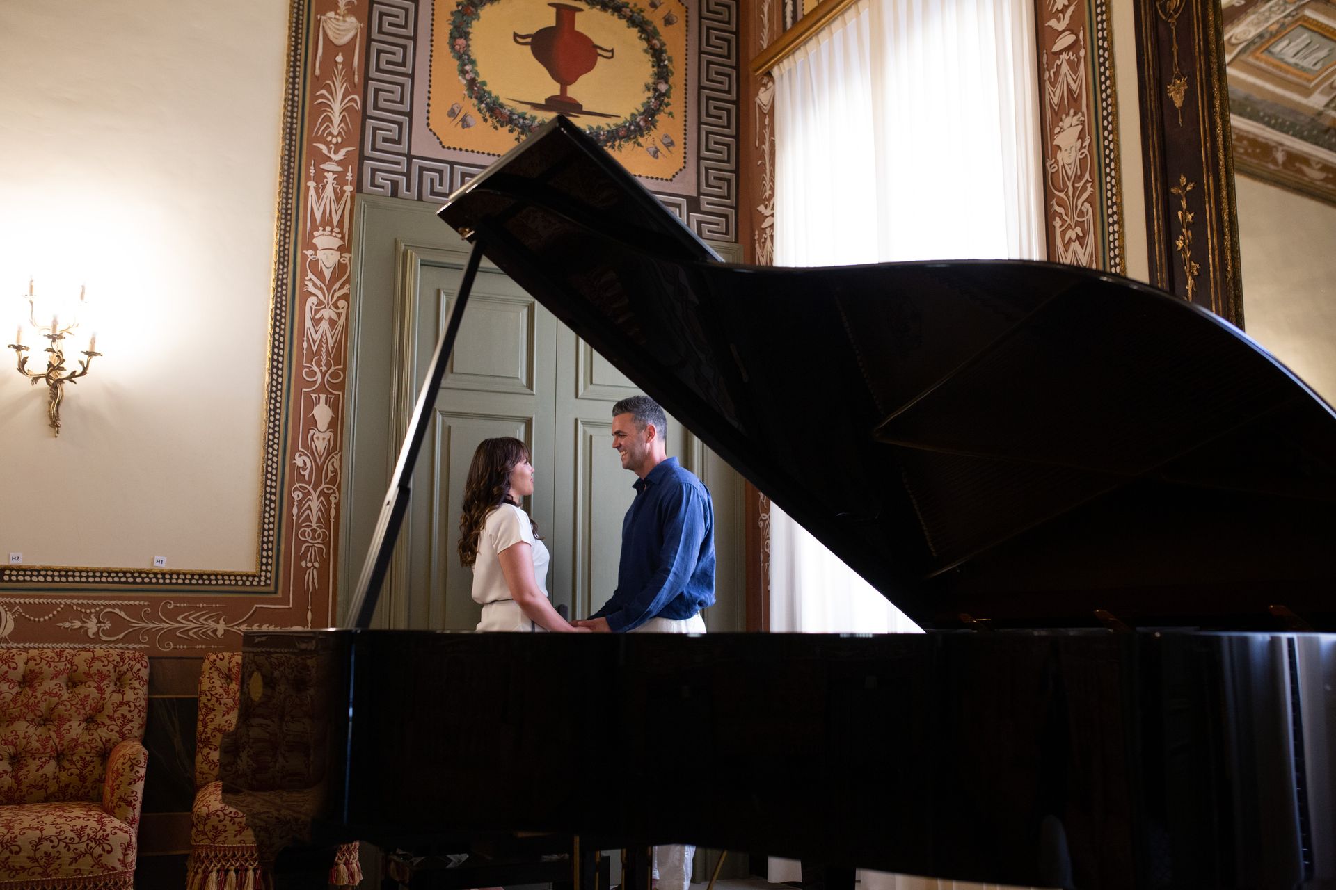 Una coppia in piedi, tenendosi per mano, vicino a un pianoforte a coda in una stanza decorata, si guarda.