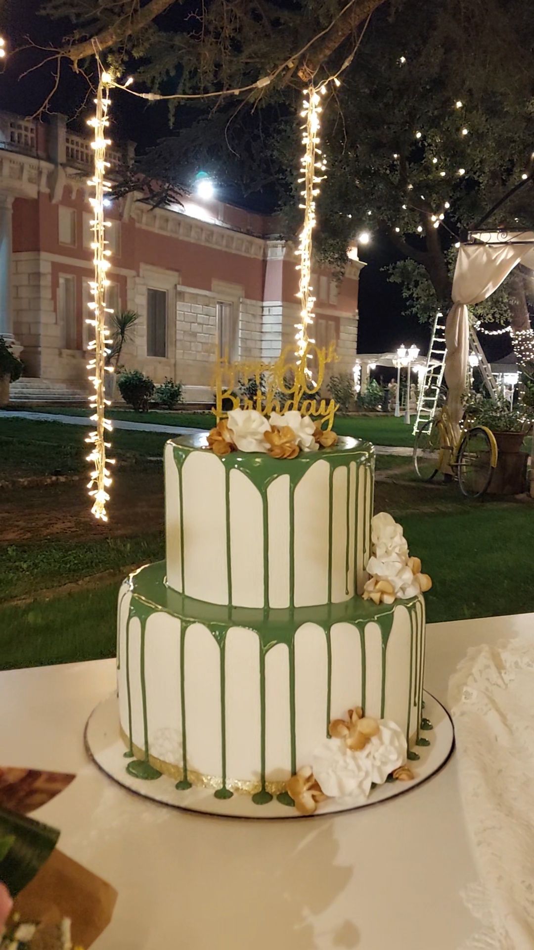 Torta bianca a due piani con gocce verdi e dettagli dorati, sormontata da un cartello con la scritta 