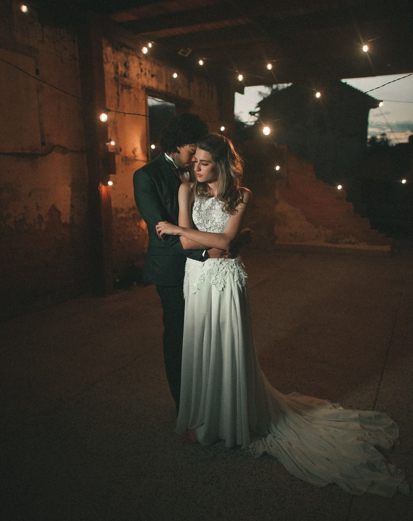 Coppia che si abbraccia in un vecchio edificio, illuminato da lucine. La sposa indossa un abito bianco, lo sposo un abito scuro.
