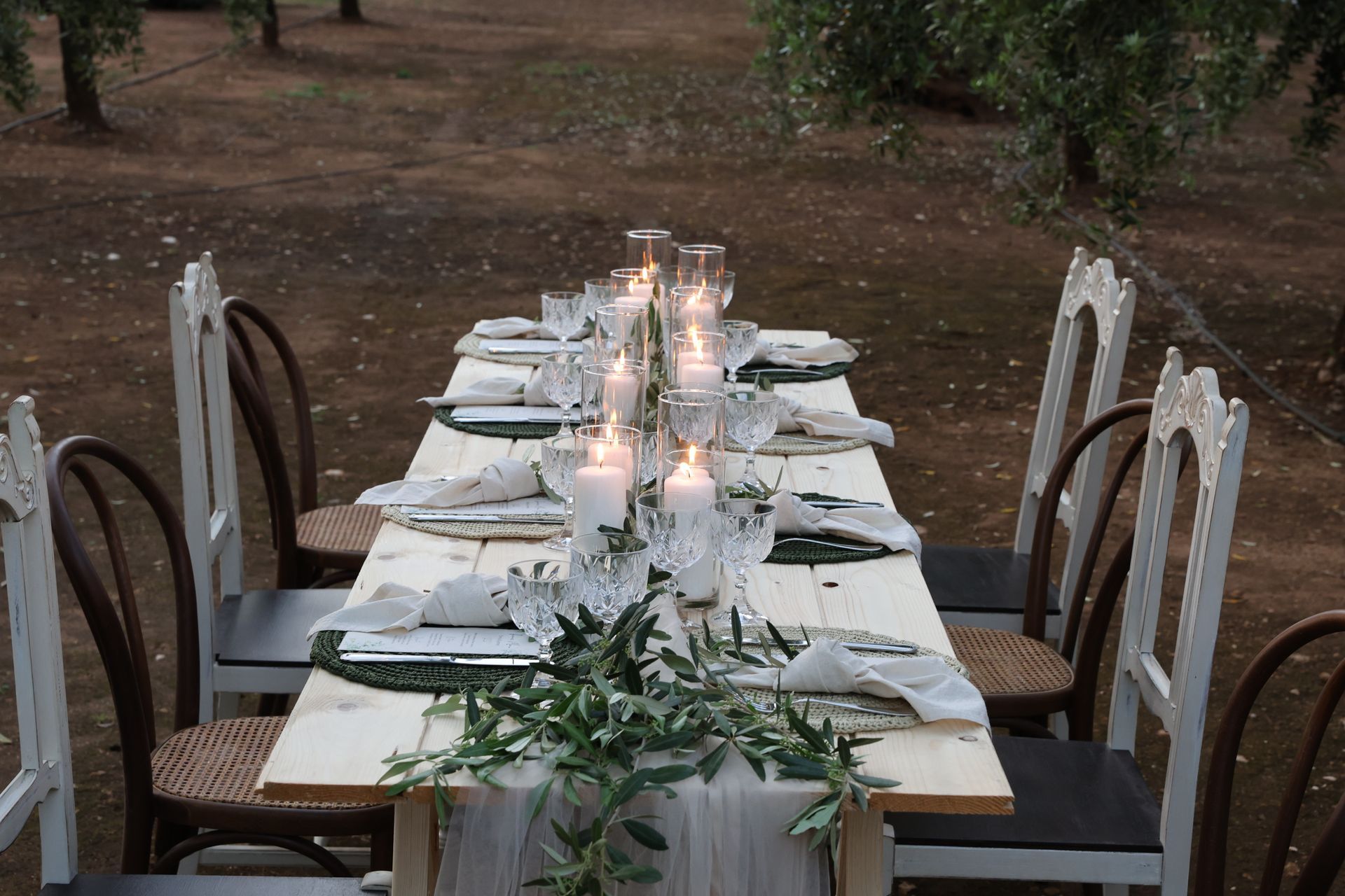 Tavolo da pranzo all'aperto apparecchiato per un pasto, con candele bianche, bicchieri e verde. Sedie marroni e bianche.