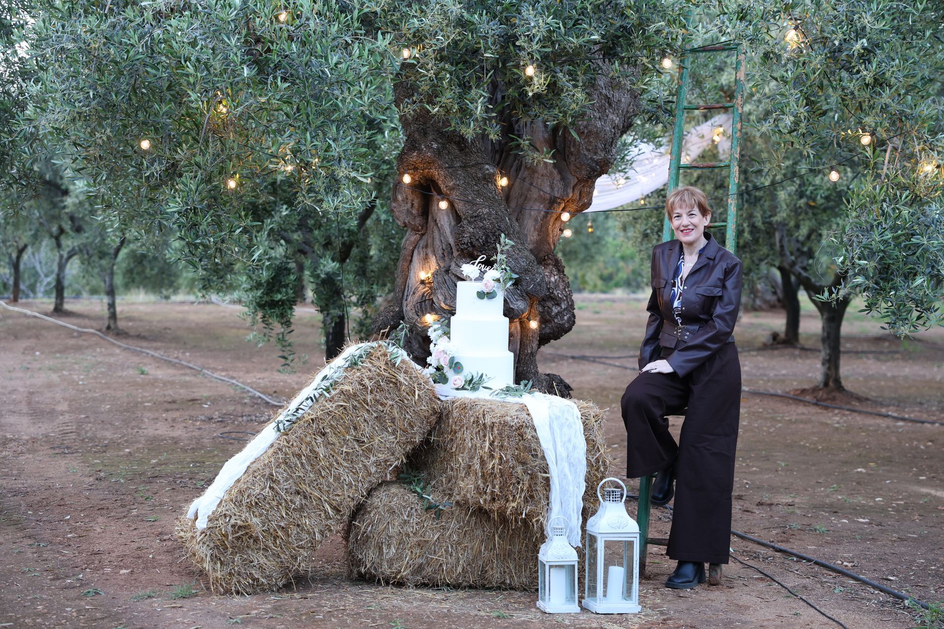 Donna accanto alla torta nuziale su balle di fieno sotto un ulivo con luci a filo.