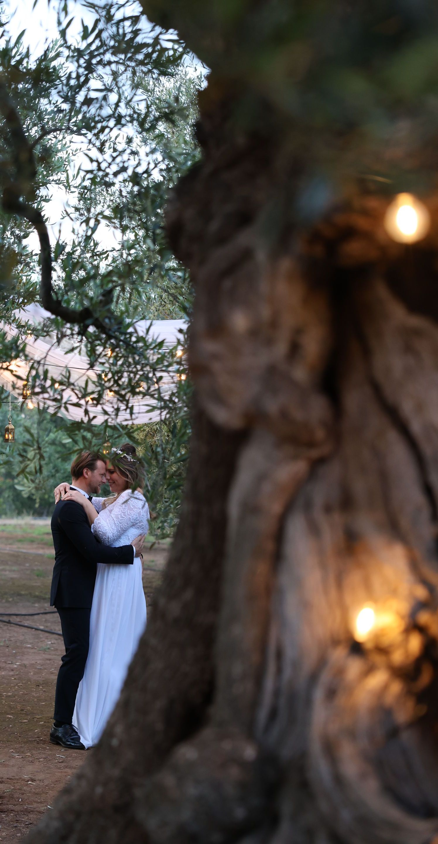 Coppia che si abbraccia in un vecchio edificio, illuminato da lucine. La sposa indossa un abito bianco, lo sposo un abito scuro.