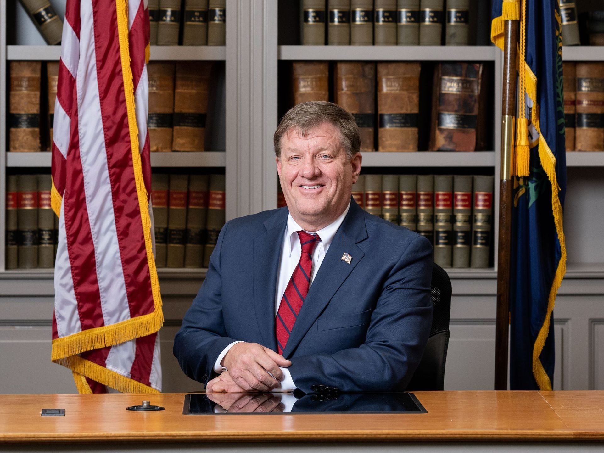 A man in a suit and tie is sitting at a desk in front of an american flag.