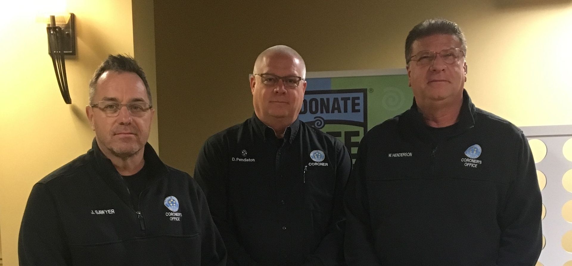 Three men are standing next to each other in front of a sign that says donate