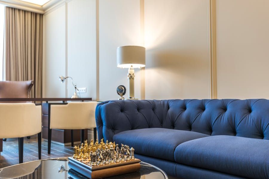 Modern living room with a navy tufted sofa, blue chair, floor lamp, and chess set on a glass coffee table
