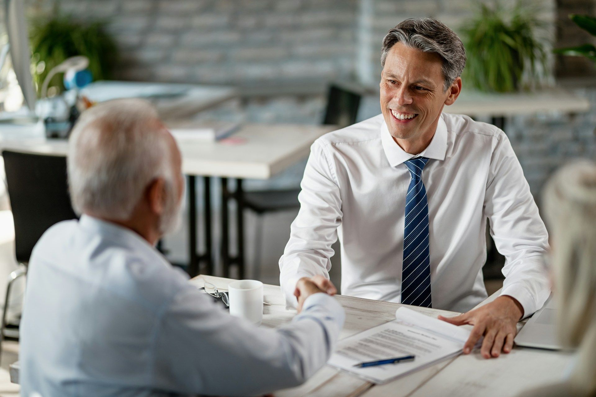 Man in tie smiles, handing papers to another man at a table in an office setting.