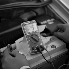 Image en noir et blanc d'un homme qui vérifie la tension d'une batterie de voiture à l'aide d'un multimètre