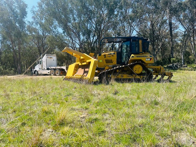 Yellow bulldozer with brush cutter on a grassy field, semi-truck in the background.