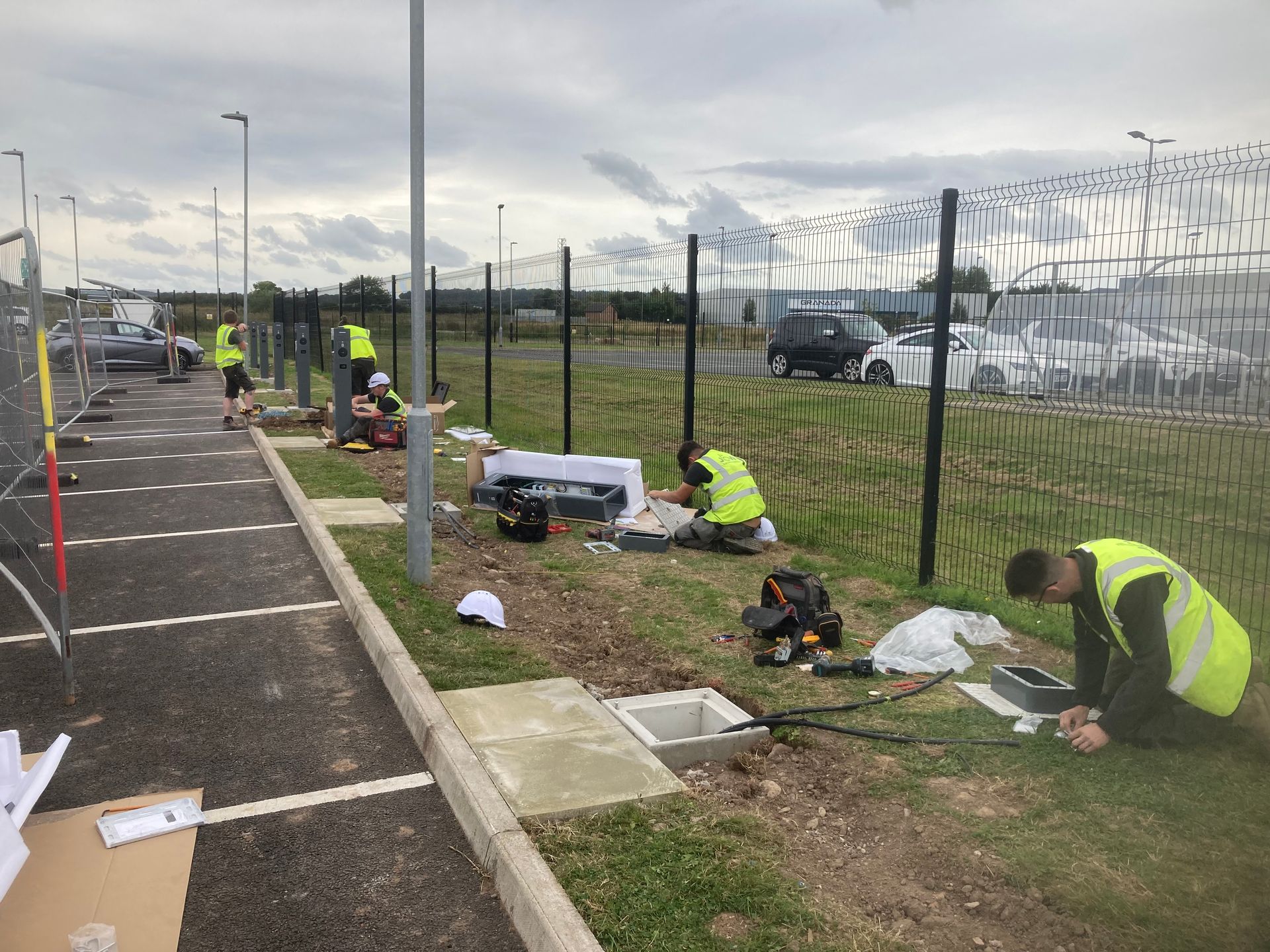 Workers in high-visibility vests perform electrical work on a grassy strip beside a paved parking area and metal fence.