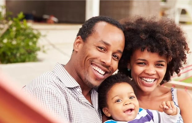 Smiling Black family of three outdoors: man, woman, and baby.