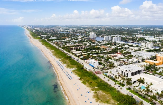Aerial view of a sunny beach with turquoise water, sandy shore, and city buildings in the background.